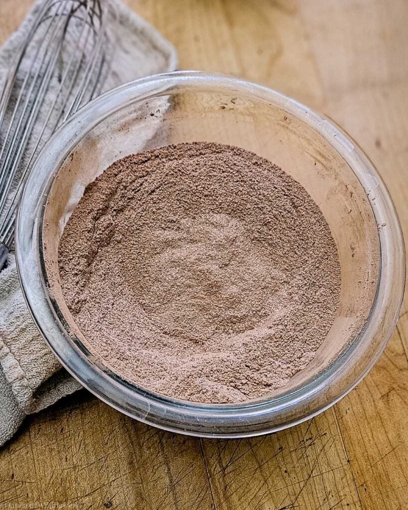 Blended brownie mix dry ingredients in a mixing bowl on the table with a linen towel & whisk.
