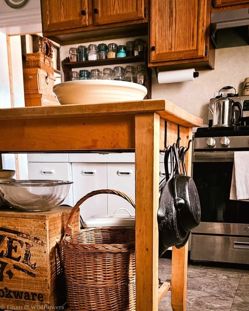 Cozy kitchen with a wooden island, hanging cast iron pans, a wicker basket, and glass jars. The stove and wooden cabinets add a warm, rustic feel.