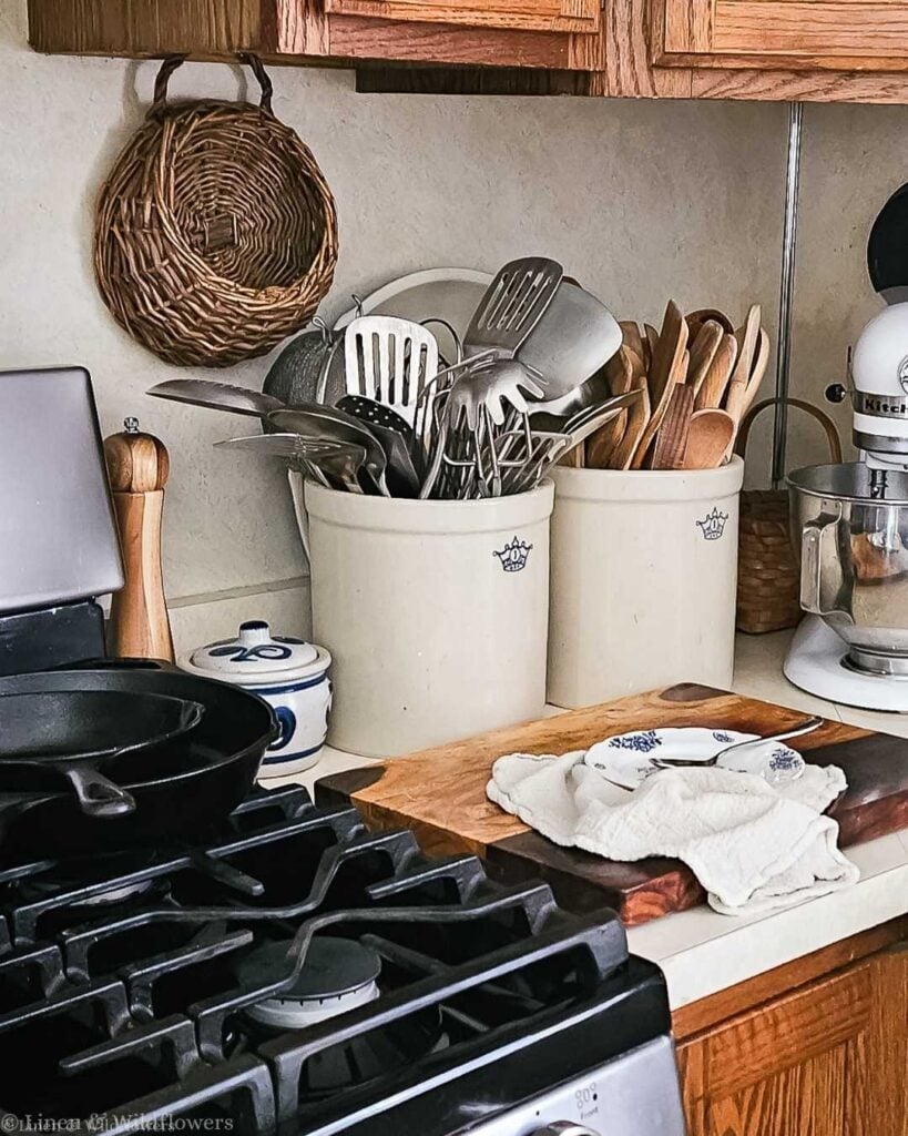 A cozy kitchen scene with utensils in ceramic jars, a cast iron skillet on a stove, a wooden cutting board, and a mixer, conveying a warm, rustic feel.