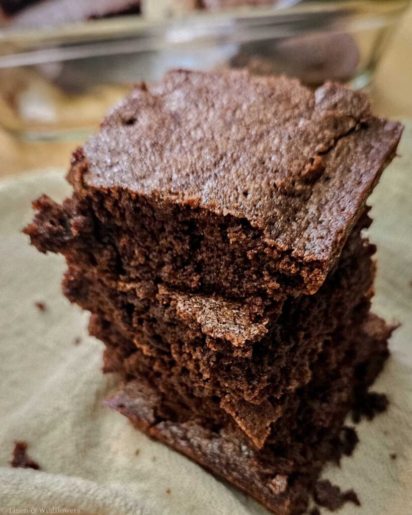A stack of three rich, fudgy brownies on a light cloth. The brownies have a slightly crumbly texture, conveying warmth and indulgence.