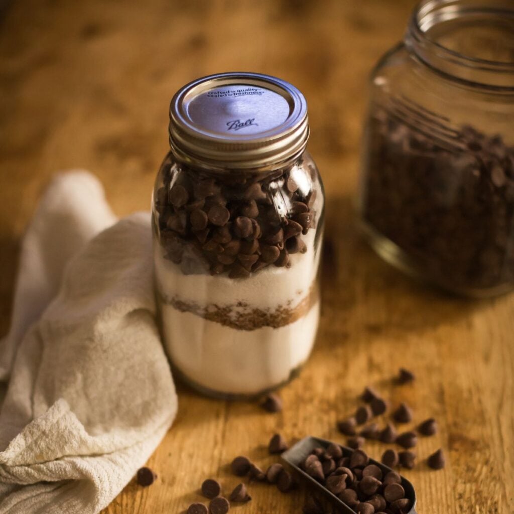 A rustic setting with a mason jar filled with layered baking ingredients, topped with chocolate chips. Nearby, a cloth and scattered chips create a cozy, homely feel.