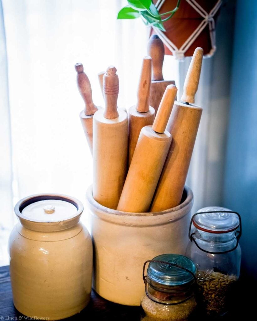A collection of wooden rolling pins in a ceramic pot beside two glass jars and a ceramic canister. Sunlight filters through sheer curtains, creating a cozy kitchen ambiance.