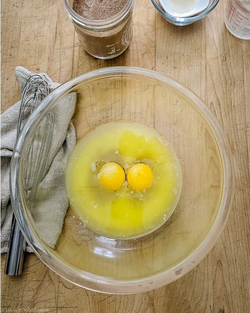 A clear bowl with cracked eggs on a wooden table. Nearby are a whisk, cloth, and jars of cocoa powder and milk, suggesting baking preparations.