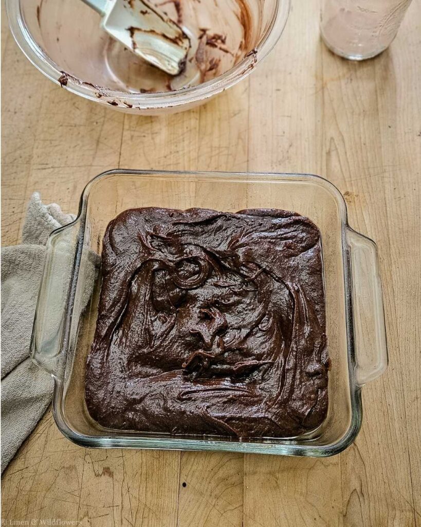 Brownie batter spread in a clear glass baking dish rests on a wooden countertop, next to a mixing bowl with chocolate remnants and a spatula. Cozy kitchen vibe.