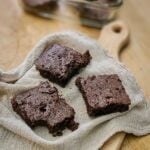 Three homemade chocolate brownies on a textured beige cloth, placed on a wooden surface. One brownie has a bite taken out, adding a cozy, inviting feel.
