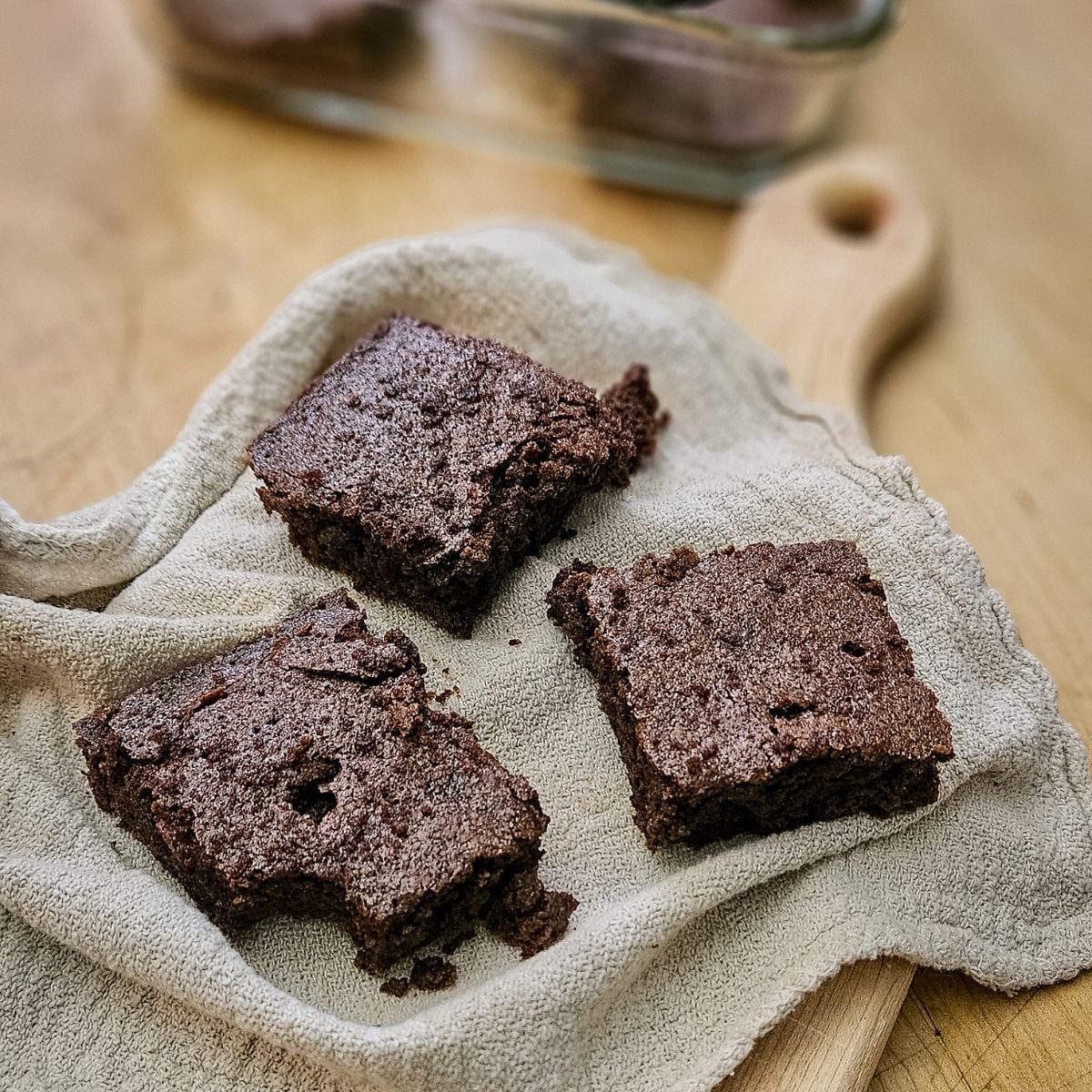 Three homemade chocolate brownies on a textured beige cloth, placed on a wooden surface. One brownie has a bite taken out, adding a cozy, inviting feel.