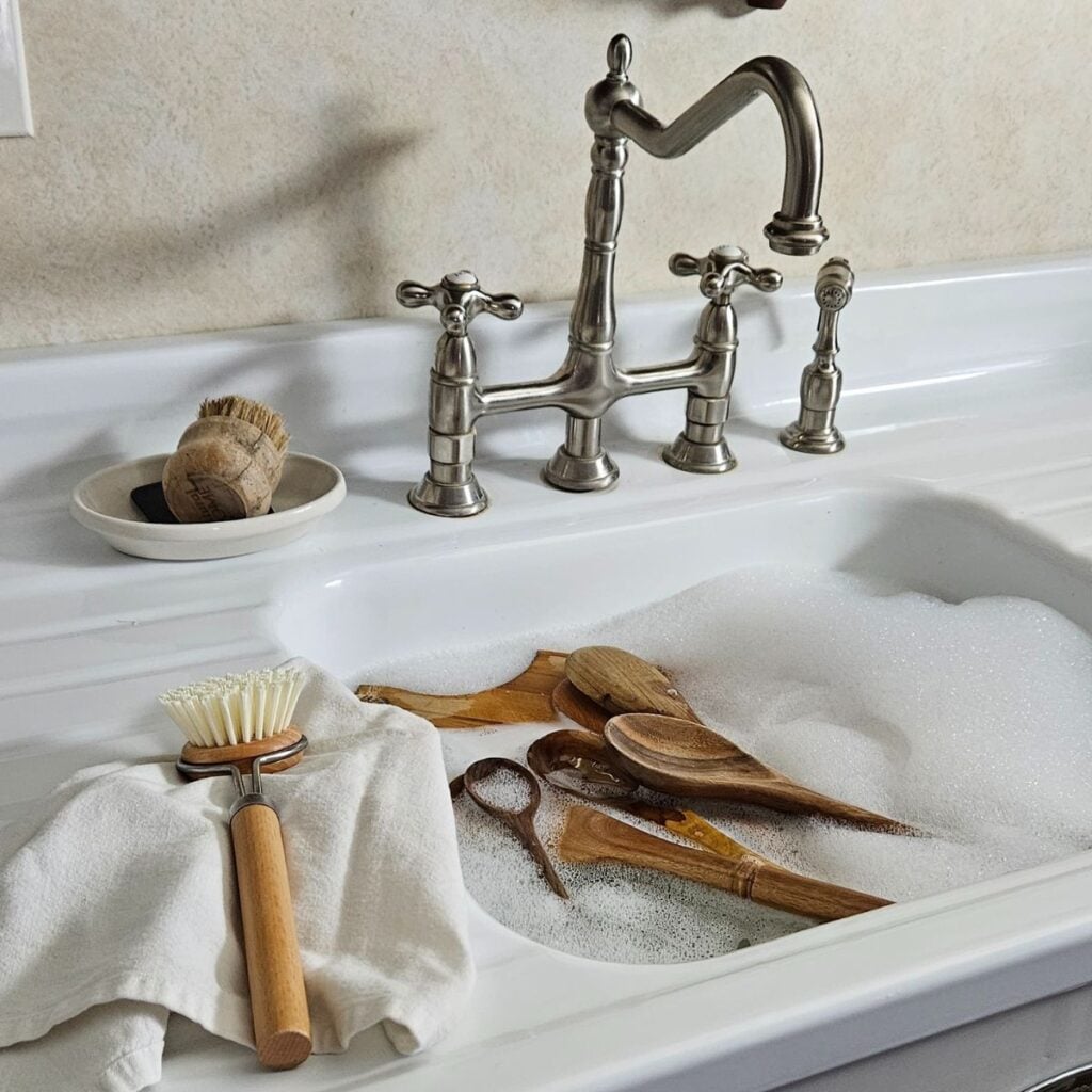 A farmhouse sink filled with soapy water and wooden utensils. A vintage-style faucet, a brush, and towel are nearby, conveying a cozy kitchen vibe.