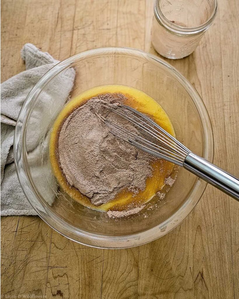 A glass bowl on a wooden table contains a mixture of chocolate powder and yellow batter, being whisked. A cloth and empty jar are nearby. Baking preparation.