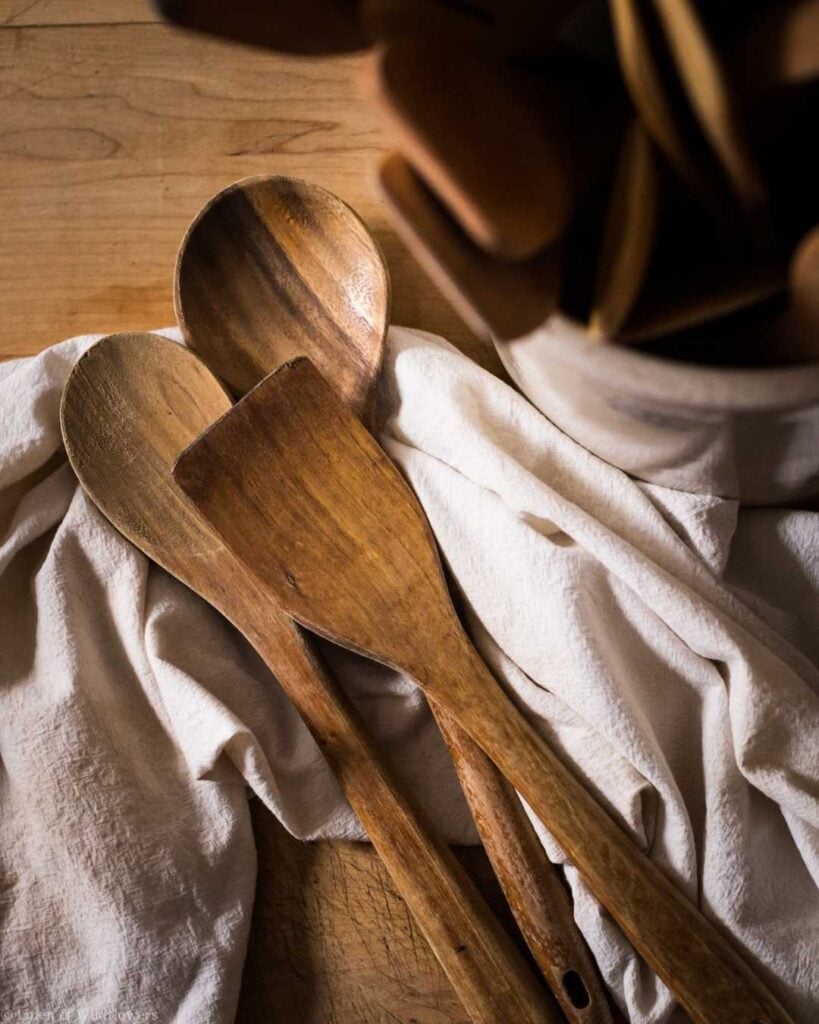 Wooden spoons rest on a rumpled white cloth beside a ceramic crock on a wooden surface, evoking a warm, rustic kitchen vibe.