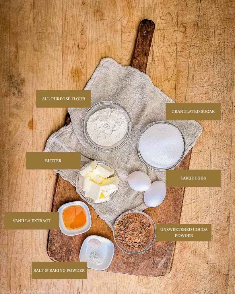 Baking ingredients labeled on a wooden board include flour, sugar, butter, eggs, cocoa, vanilla extract, and salt. Rustic kitchen setting with a cozy vibe.