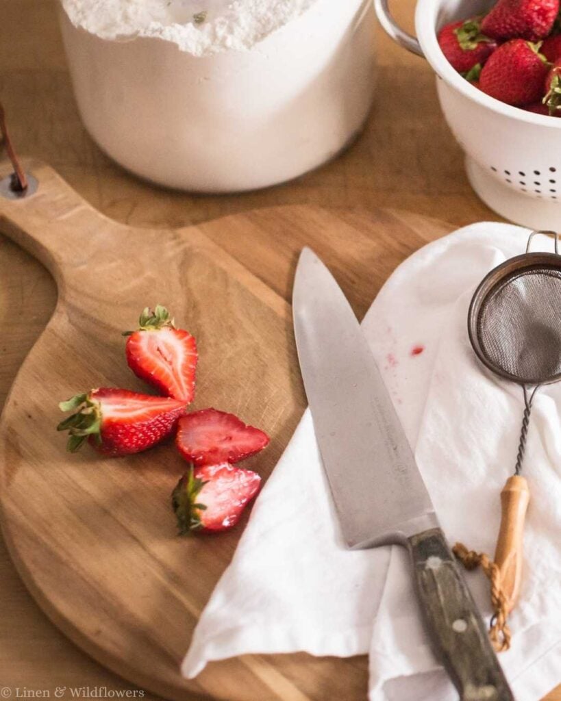 Freshly sliced strawberries sit on a wooden board next to a knife, a sifter, and a white cloth. A pot of flour and a colander of strawberries are nearby.