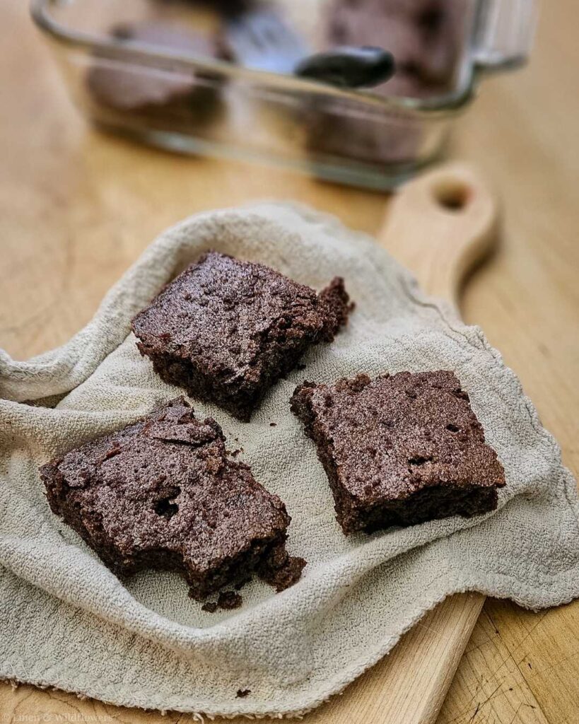 Three homemade brownies sit on a cloth over a wooden board, with a baking dish in the background. The scene conveys a cozy, inviting atmosphere.