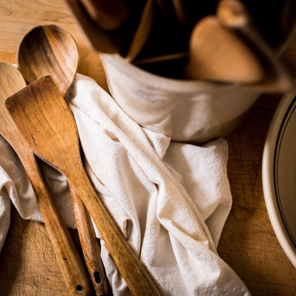 Wooden spoons rest on a rumpled white cloth beside a ceramic crock on a wooden surface, evoking a warm, rustic kitchen vibe.
