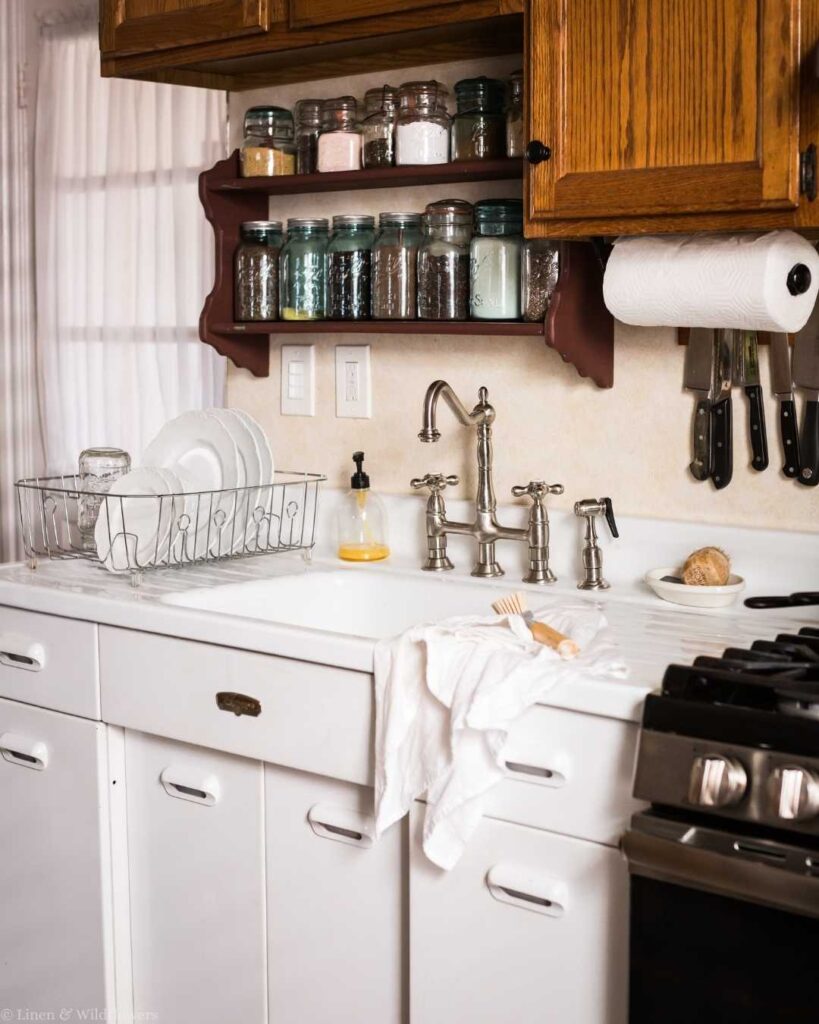 Cozy kitchen with wooden cabinets and open shelves holding jars. A dish rack sits on a white sink with a vintage faucet and dish soap.