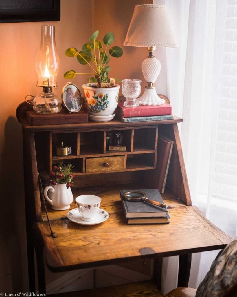 Cozy vintage desk with a warm lamp, potted plant, and vintage decor. A tea set, books, and magnifying glass sit on the wooden surface, evoking nostalgia.