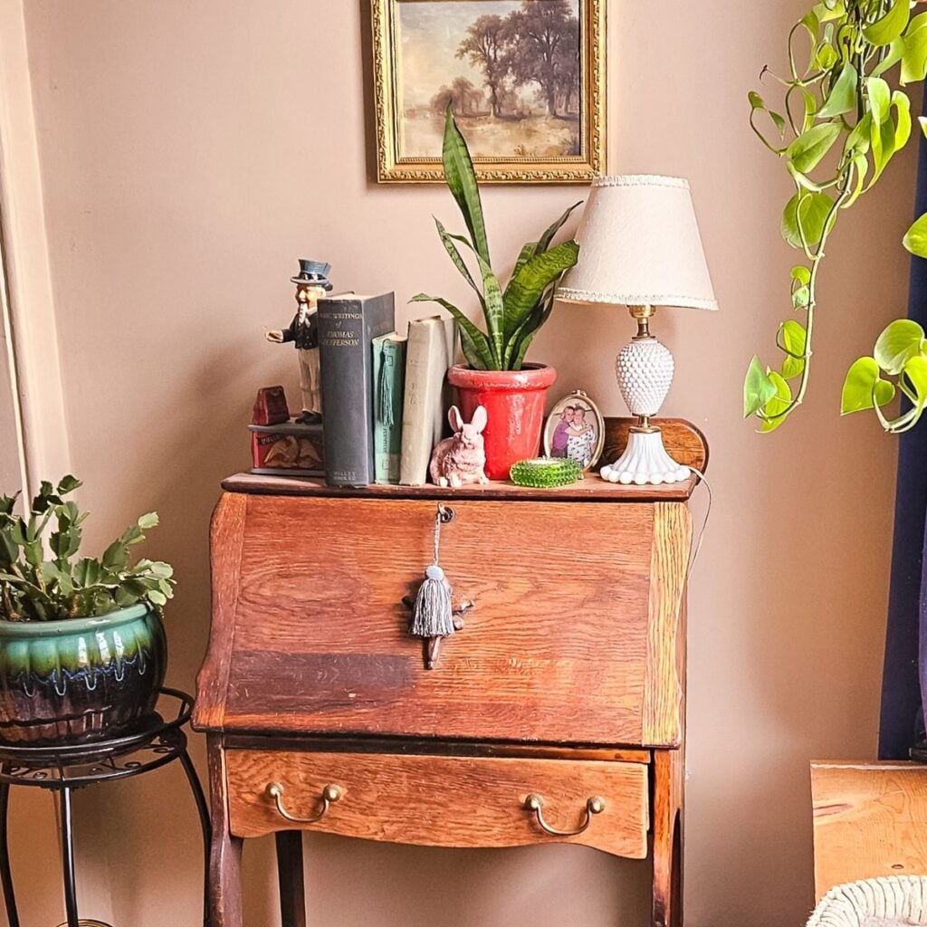 A cozy interior with a vintage wooden desk adorned with books, a potted plant, a small lamp, and decorative items. A painting with trees hangs above.