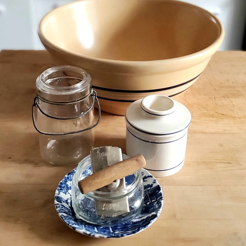 A rustic kitchen scene with a large beige mixing bowl, glass mason jar, vintage blue-patterned dish with soap bars, and a ceramic container on wood table.
