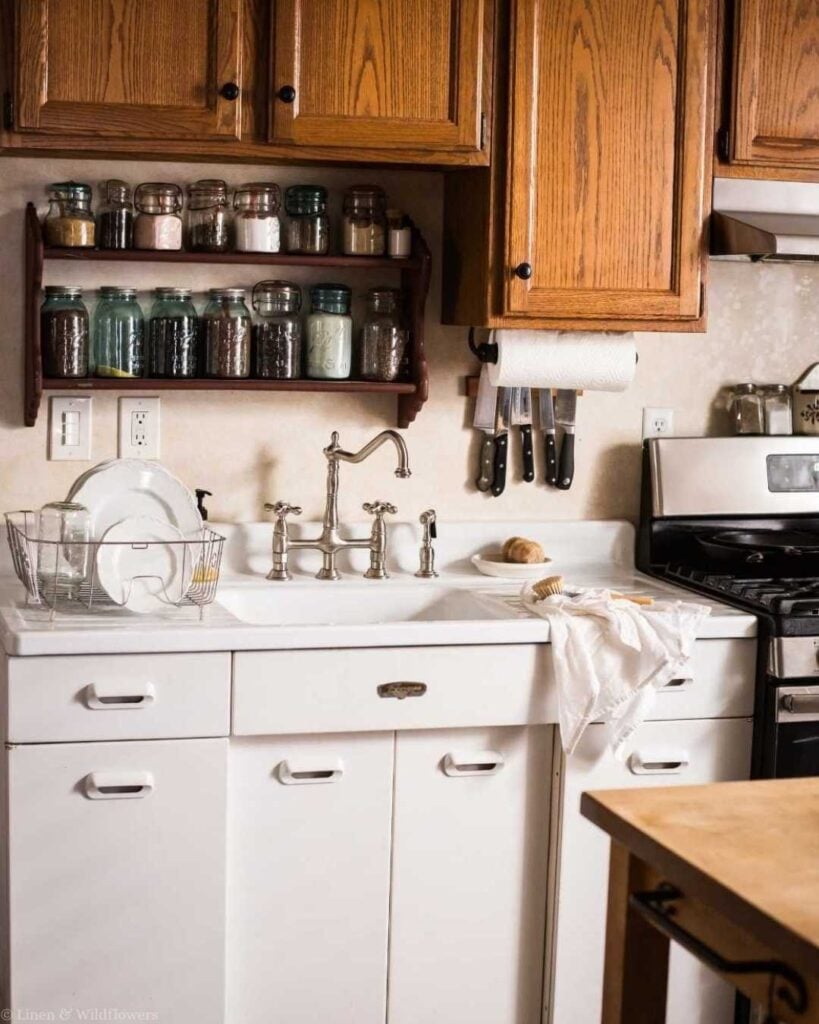 Cozy kitchen scene with rustic wooden cabinets, a white farmhouse sink, vintage jars on a shelf, and dishware drying. Warm and inviting atmosphere.
