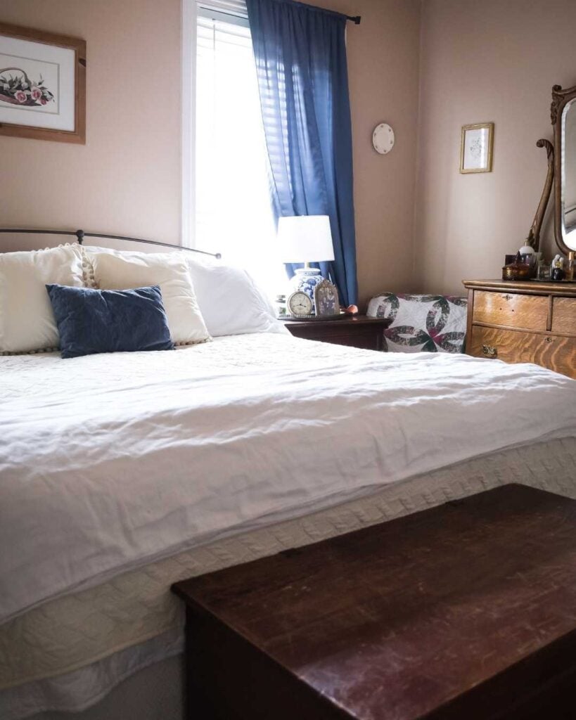 Cozy bedroom with a large bed, blue pillow, white bedding, and wooden dresser. Soft lighting from a window with blue curtains creates a serene atmosphere.