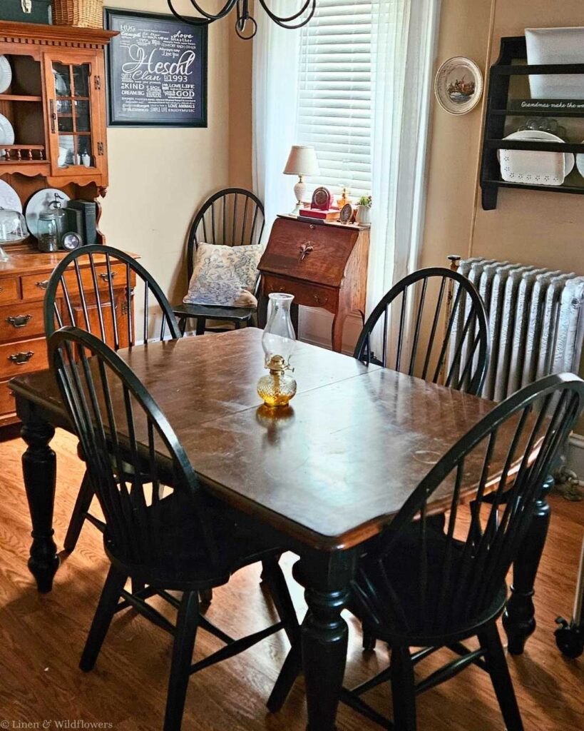 Cozy dining room with a wooden table set, six chairs, vintage hutch, framed art, and sunlight streaming through blinds. Warm, inviting atmosphere.