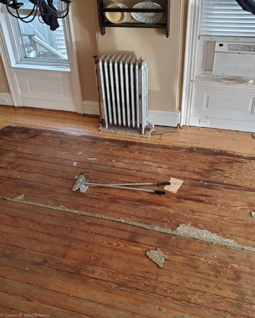 Wooden floor with damaged patches and tools scattered, indicating renovation. A radiator and shelves with plates are against a beige wall, giving a rustic feel.