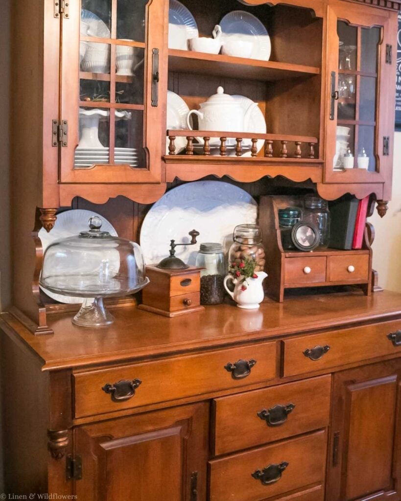 A wooden china cabinet displays white dishes, a glass cake stand, jars, a vintage scale, and a small drawer set, creating a cozy, rustic ambiance.