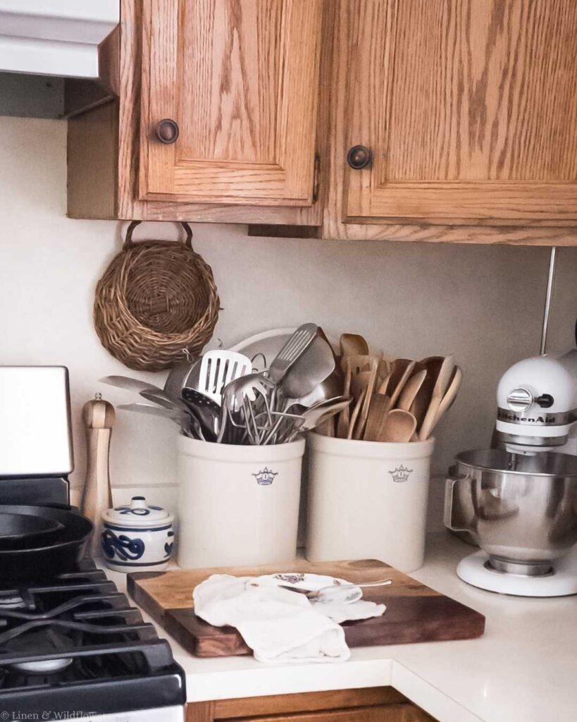 Cozy kitchen corner with wooden cabinets, utensils in ceramic pots, a wicker basket, a cutting board with cloth, and a mixer. Warm, rustic ambiance.