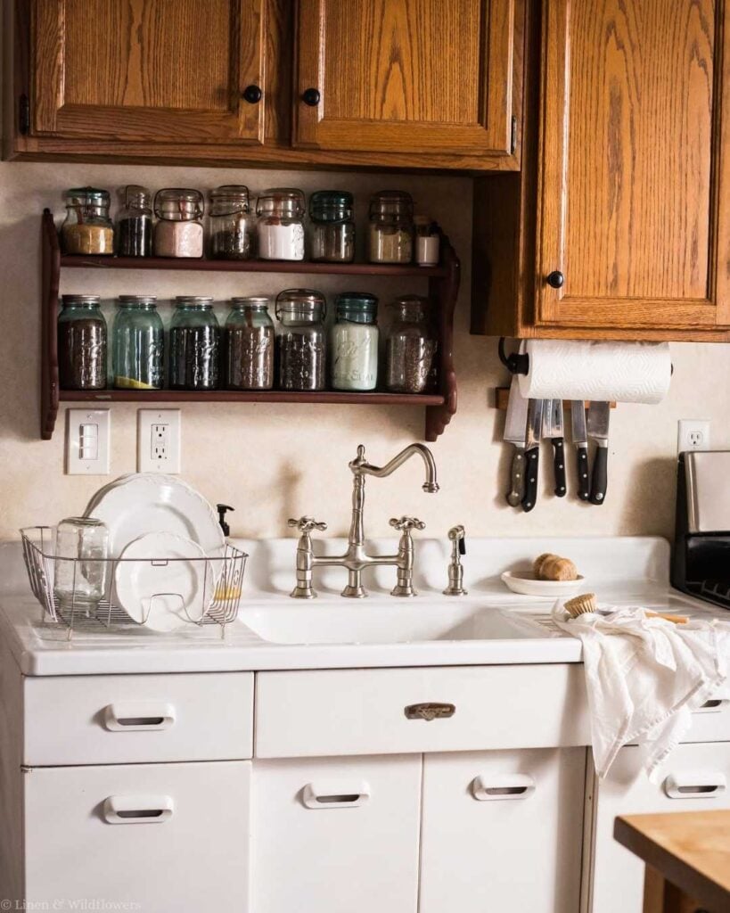 Cozy kitchen with a white sink, vintage faucet, and wooden cabinets. Shelves hold jars of spices. A cloth and stacked dishes rest on the counter.