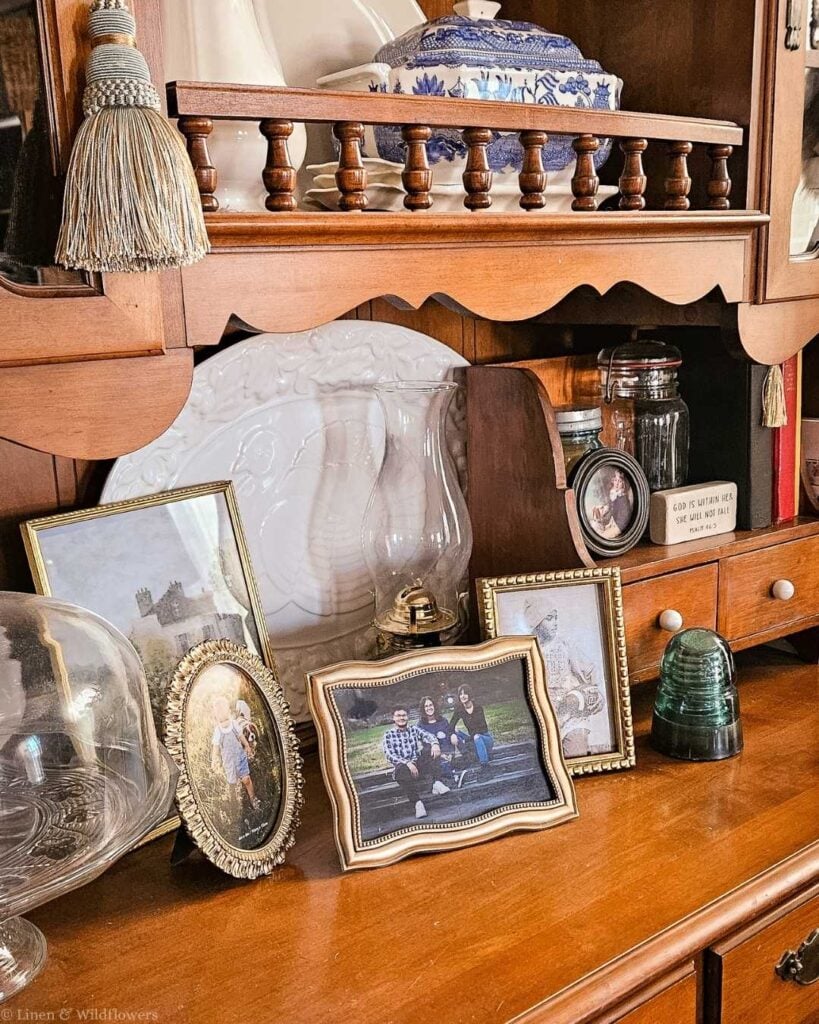 A wooden shelf displays family photos in ornate frames, china, and glass jars, creating a nostalgic, homely atmosphere. The tone is warm and sentimental.