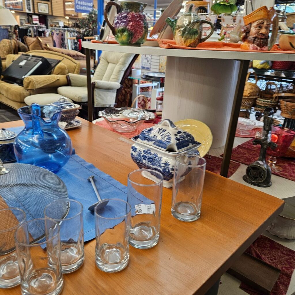 Thrift store interior with assorted glassware and ceramics. A blue pitcher, floral jug, and blue-patterned dishes are displayed on wooden shelves.