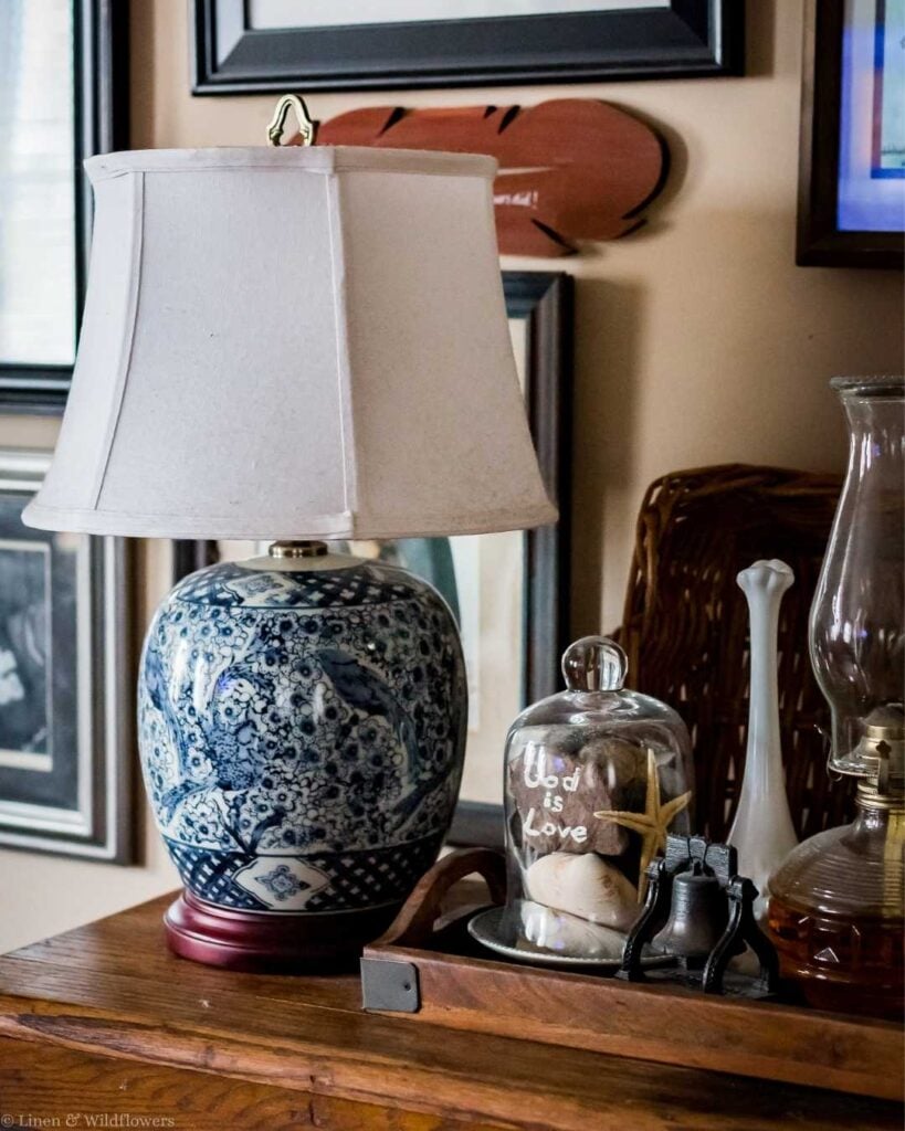 Elegant room decor featuring a blue and white ceramic lamp with a white shade. Nearby, a glass cloche encloses shells and a starfish, labeled "God is Love." An oil lamp and framed art add a cozy, nostalgic feel.