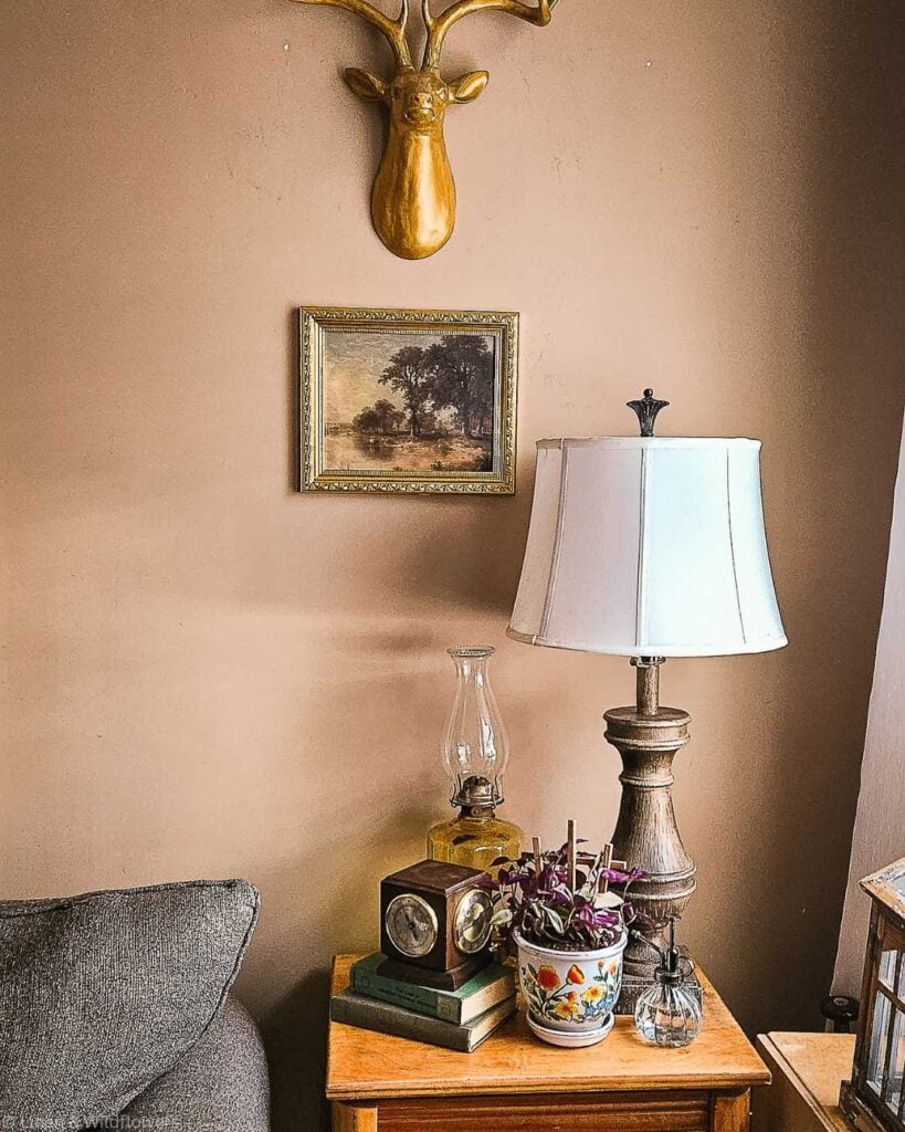 Cozy living room corner with a wooden table holding a lamp, potted plant, clock, and books. A deer head sculpture and landscape painting adorn the tan wall.