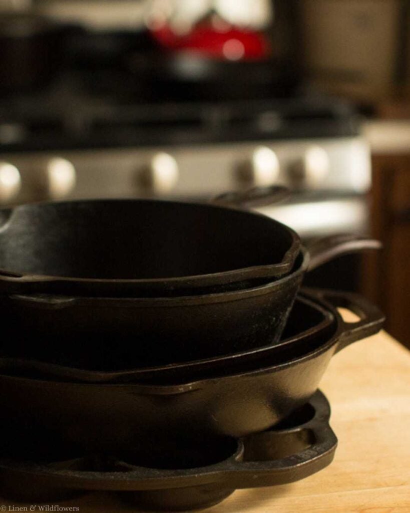 Stacked black cast iron skillets on a wooden countertop, with a stove and blurred red teapot in the background, creating a cozy kitchen vibe.