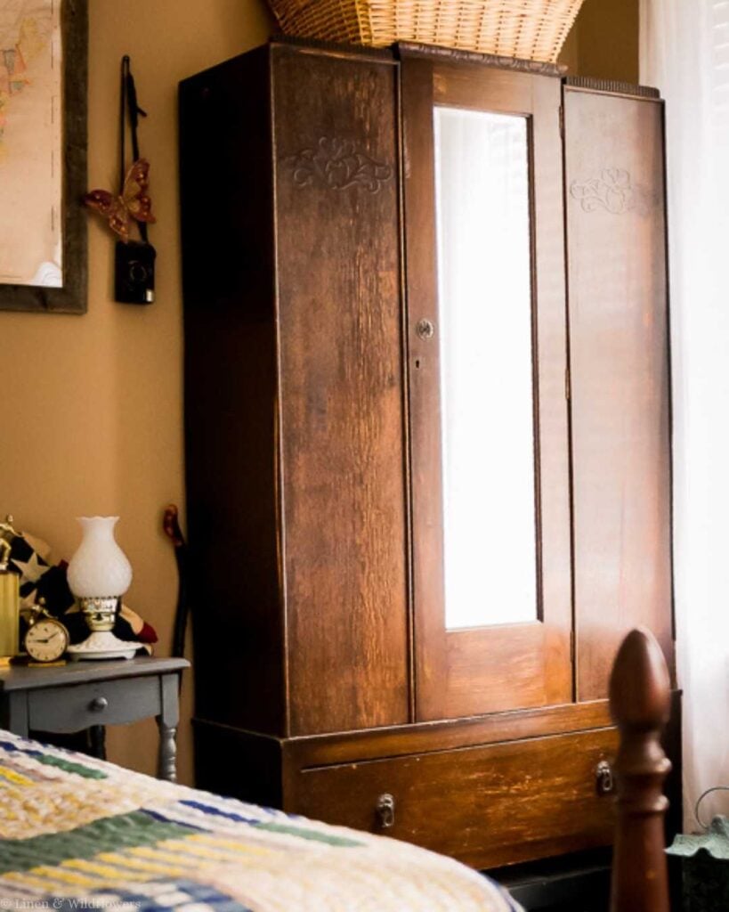A cozy bedroom with a vintage wooden wardrobe featuring carved details and a central mirror. A quilted bedspread, a bedside table with a lamp, and a basket on top of the wardrobe add warmth to the space. Natural light filters through the window blinds, creating a peaceful atmosphere.