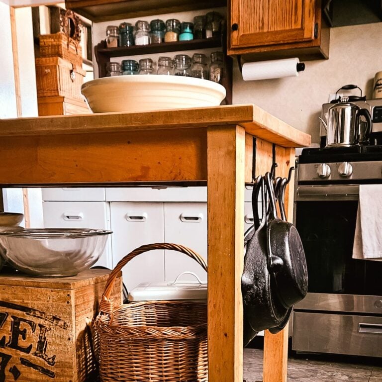 Cozy kitchen with wooden island holding bowls and cast iron pans. Shelves with spice jars, wicker basket, vintage feel. Stove and cabinets in background.