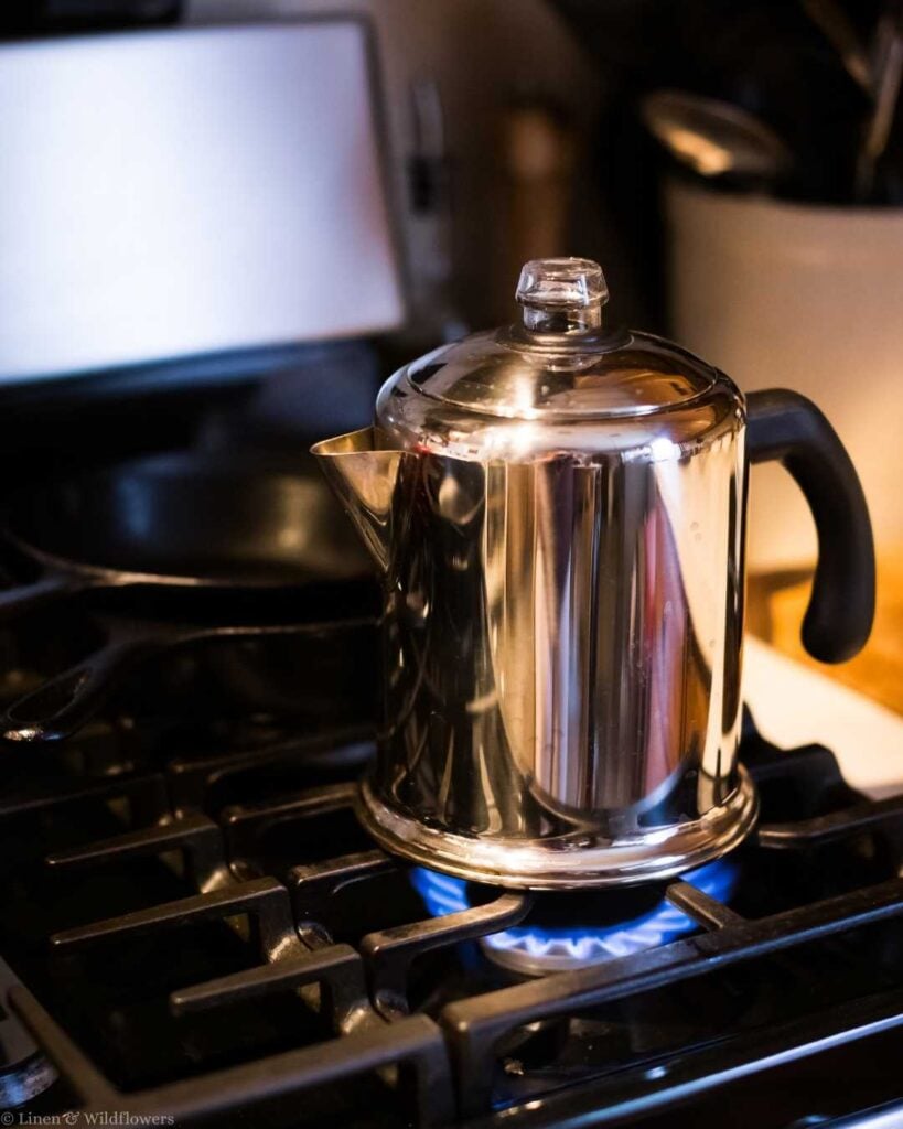 A shiny metal coffee percolator with a black handle sits on a gas stove, with blue flame visible underneath, creating a warm, cozy atmosphere.