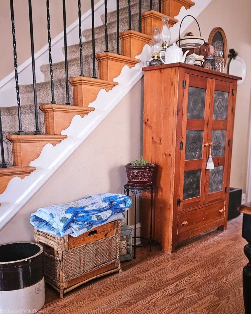 A cozy dining room with a wooden cabinet by the stairs, topped with decorative glassware. A wicker bench with a blue quilt sits beside a plant stand.