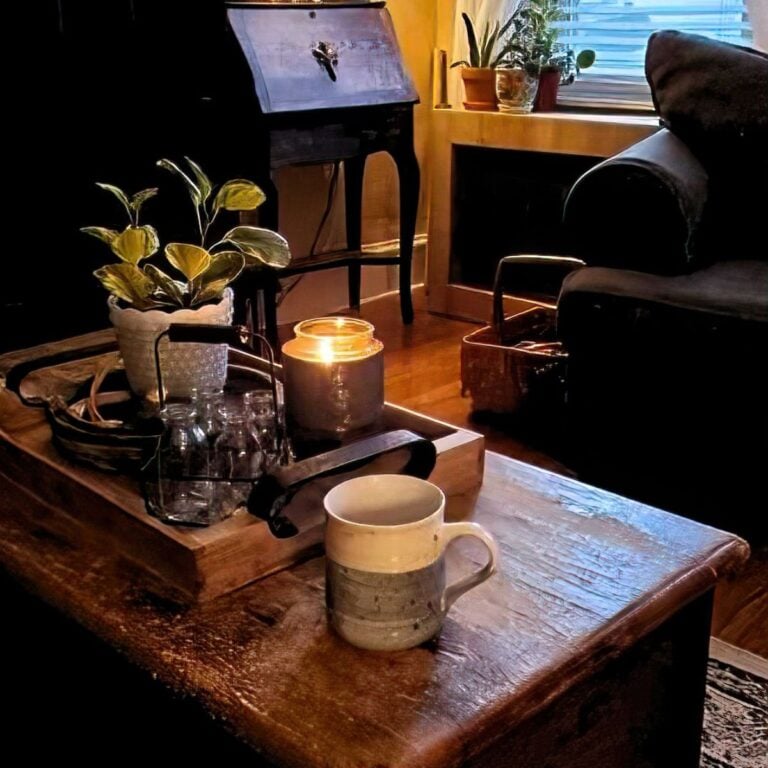 Cozy living room with a rustic wooden coffee table holding a steaming mug, glowing candle, potted plant, and glass bottles. Warm, inviting atmosphere.