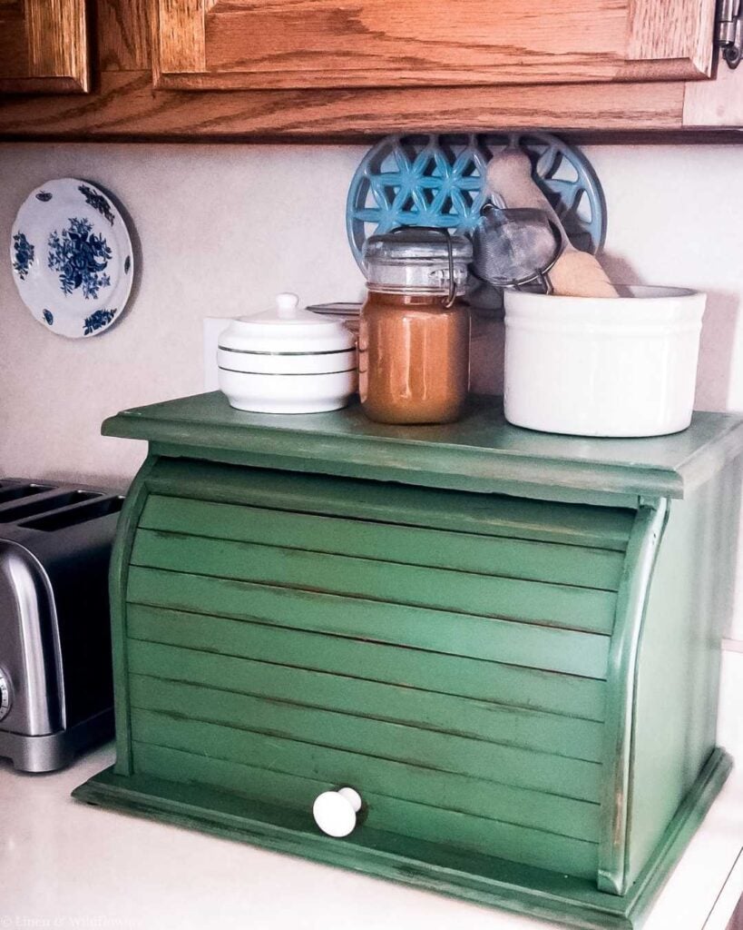 Green bread box with a white knob on a kitchen counter. A jar and ceramic containers are on top, and a blue decorative plate hangs on the wall.