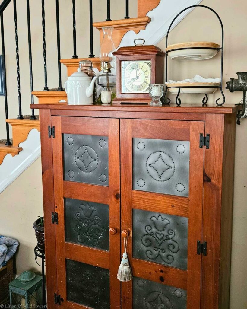 Antique punched-tin pie safe cabinet in warm cherry wood displayed near a staircase, topped with a mantel clock, white ceramic teapot, oil lamp, and a tiered wire plate stand — classic farmhouse storage and decor on a budget