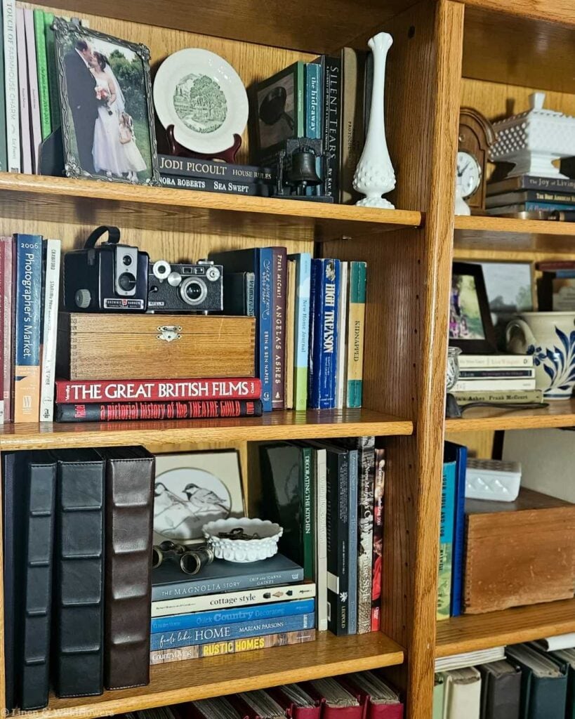 Close-up of bookcase shelves styled with vintage cameras, framed wedding photo, milk glass vase, and stacked books, including The Great British Films, in a farmhouse living room.