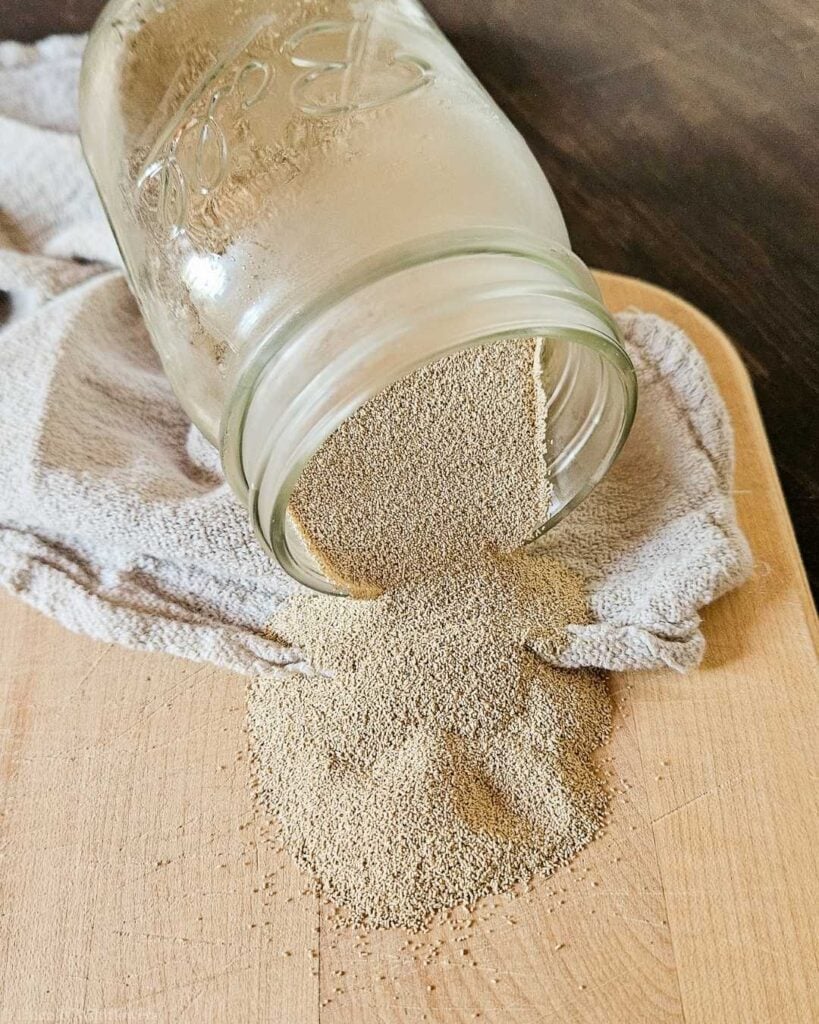 Active dry yeast granules spilling out of a glass mason jar onto a wooden cutting board, showing how to store yeast in bulk at home