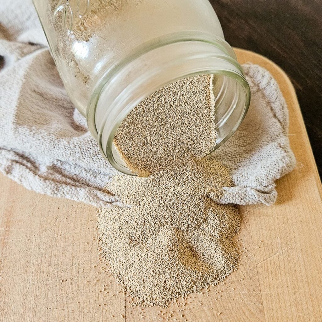 Active dry yeast granules spilling out of a tipped glass mason jar onto a wooden cutting board, illustrating how to test and use expired yeast for bread baking.
