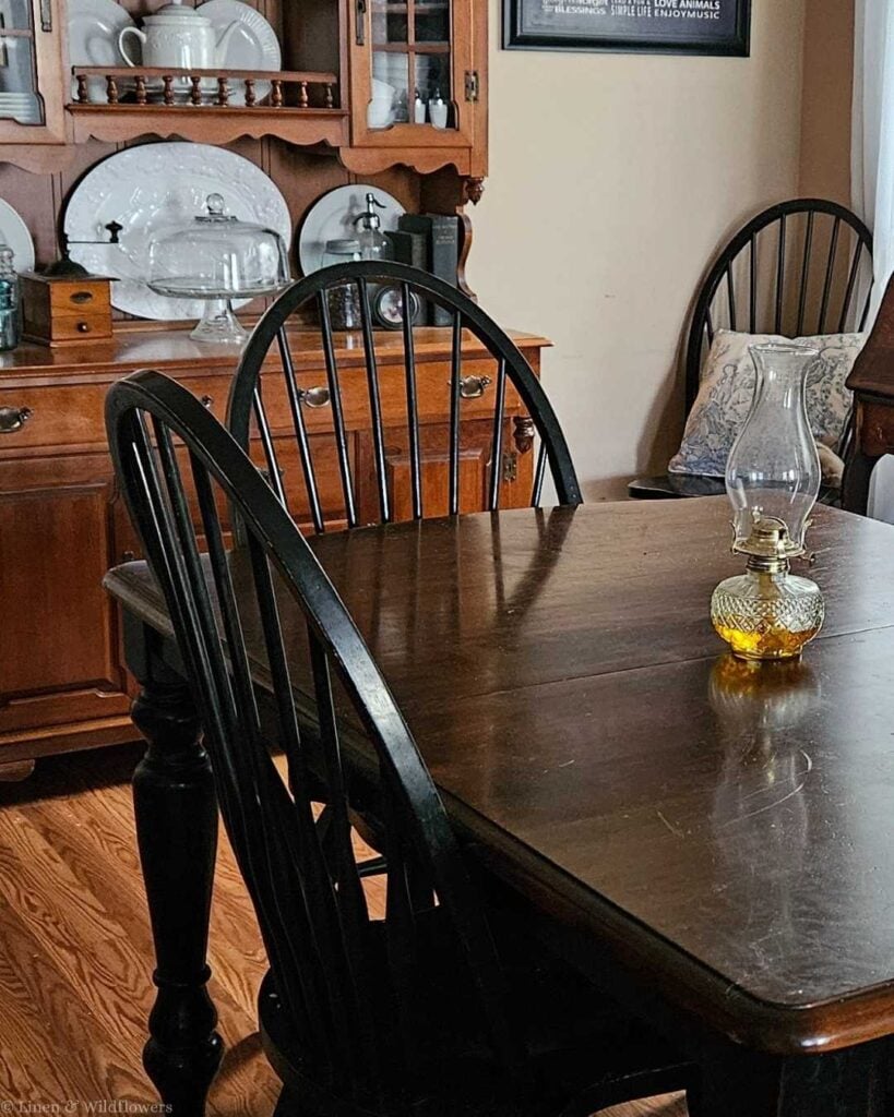 Cozy farmhouse-style dining room with dark wood drop-leaf table, black Windsor spindle chairs, warm oak china hutch displaying white ironstone dishes, and a small amber glass oil lamp centerpiece — vintage cottage dining room decor on a budget
