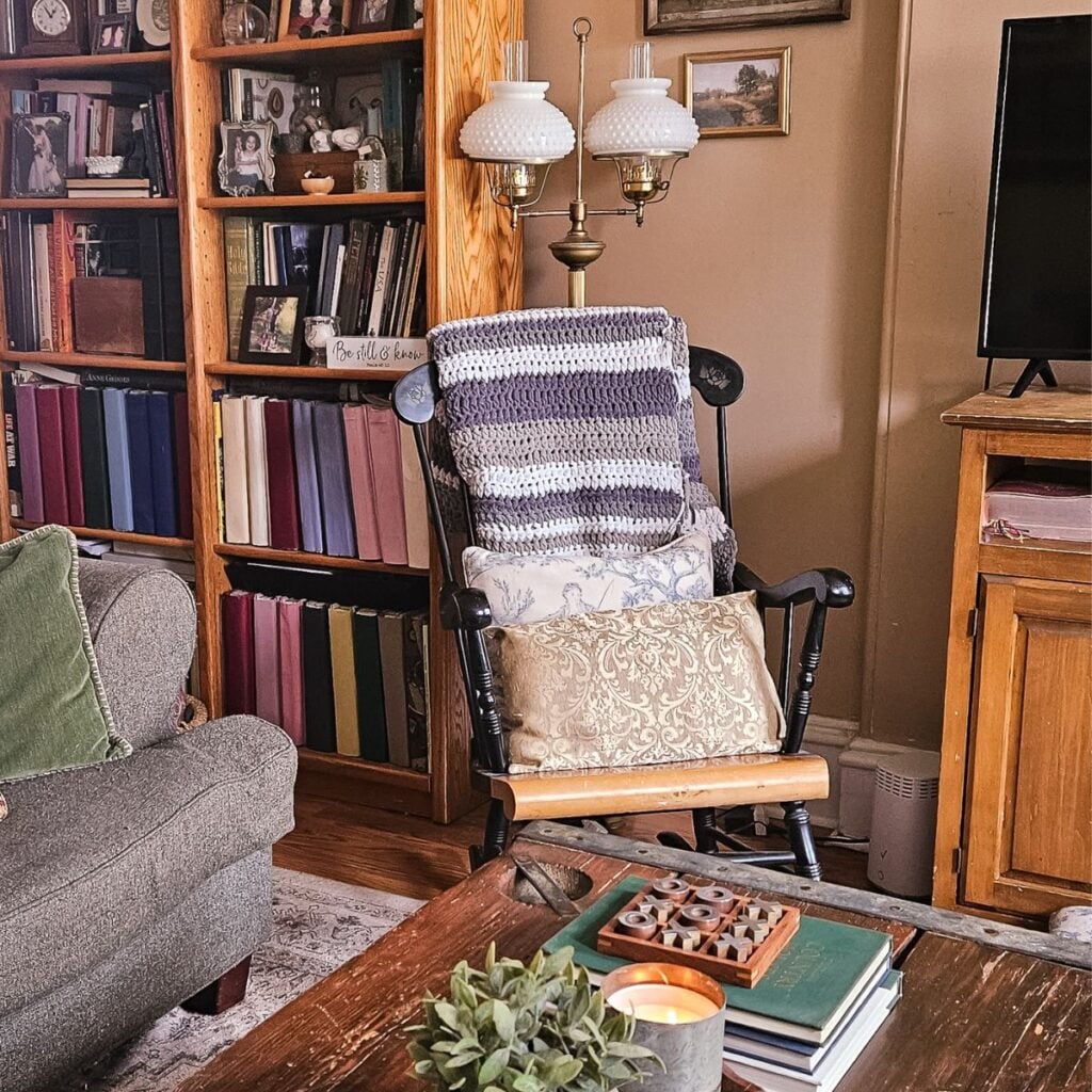 Warm and cozy living room with a black rocking chair draped in a handmade striped crochet throw, surrounded by a towering wooden bookshelf filled with books and family photos, a vintage brass hobnail milk glass floor lamp, and a rustic coffee table with candle and greenery — how to create a cozy home on a budget