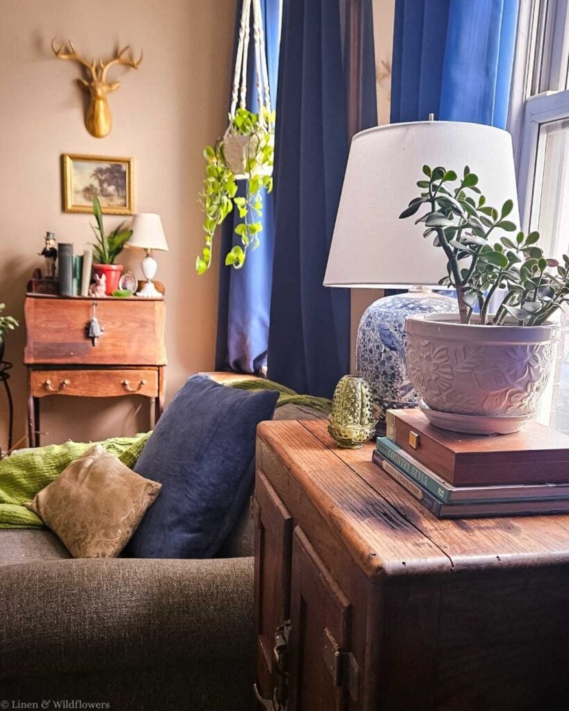 Cozy English-style living room corner with antique wooden trunk used as a side table, blue and white ginger jar lamp, jade plant in a ceramic pot, blue velvet curtains, and a hanging pothos plant — budget-friendly vintage home decor ideas