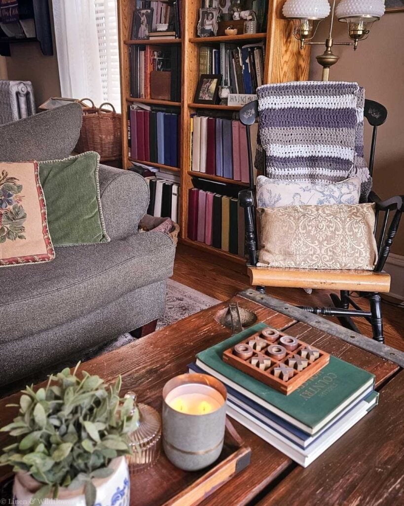 Cozy living room reading nook with floor-to-ceiling wood bookshelf filled with books, black rocking chair with crochet throw and pillows, rustic wood coffee table styled with stacked books, a lit candle, and a tic-tac-toe board — affordable cozy home decor on a budget