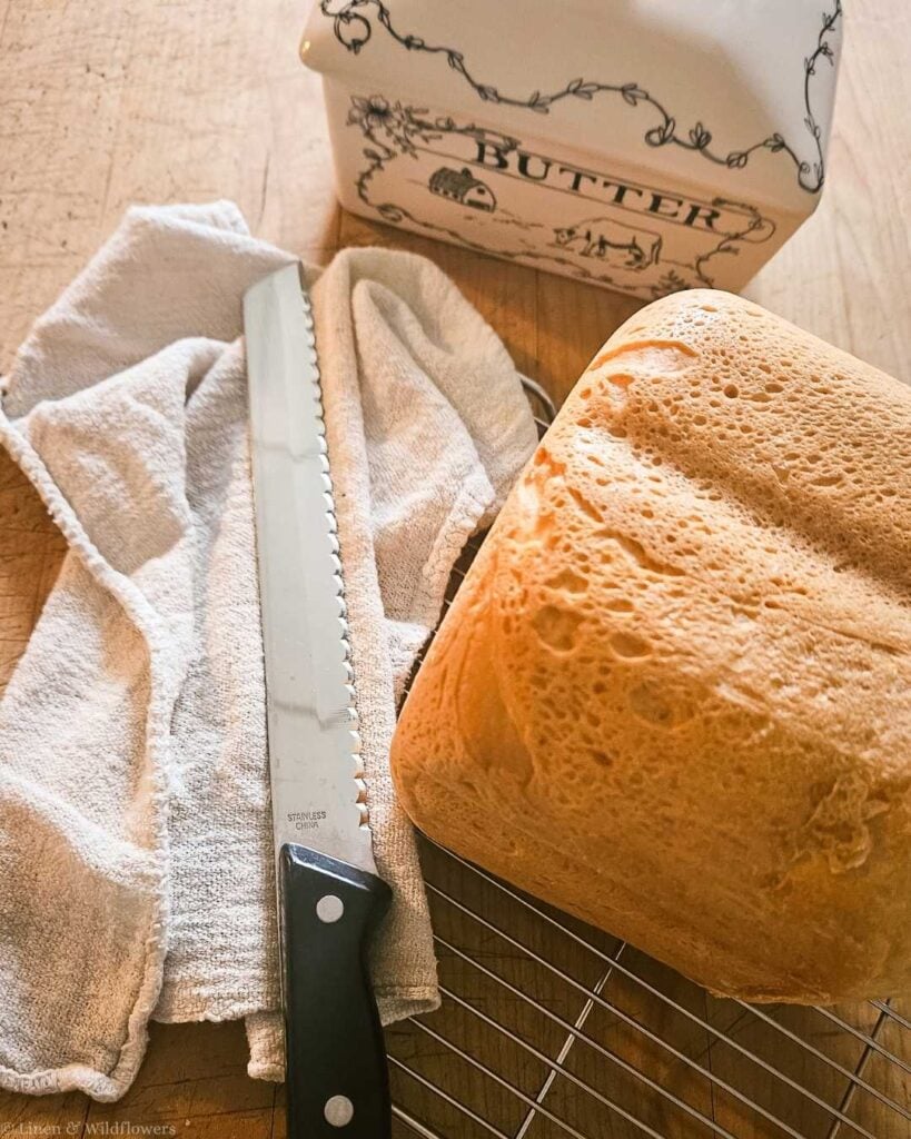 Golden homemade sandwich bread loaf cooling on a wire rack next to a bread knife and butter dish, successfully baked using proofed expired yeast.