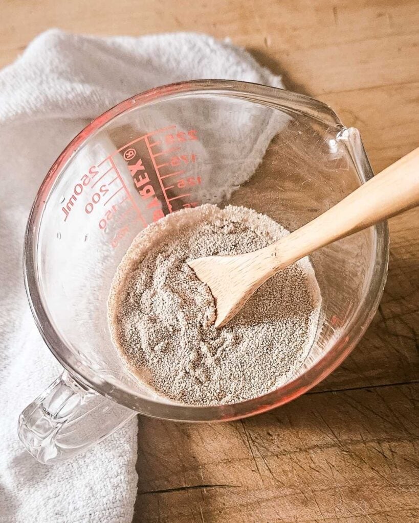 Dry active yeast granules being stirred into warm water in a glass measuring cup with a wooden spoon to test if expired yeast is still active.
