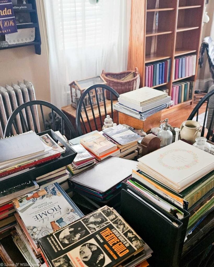 Books and collectibles spread across a dining table are being sorted and organized before being styled back onto an empty bookcase.